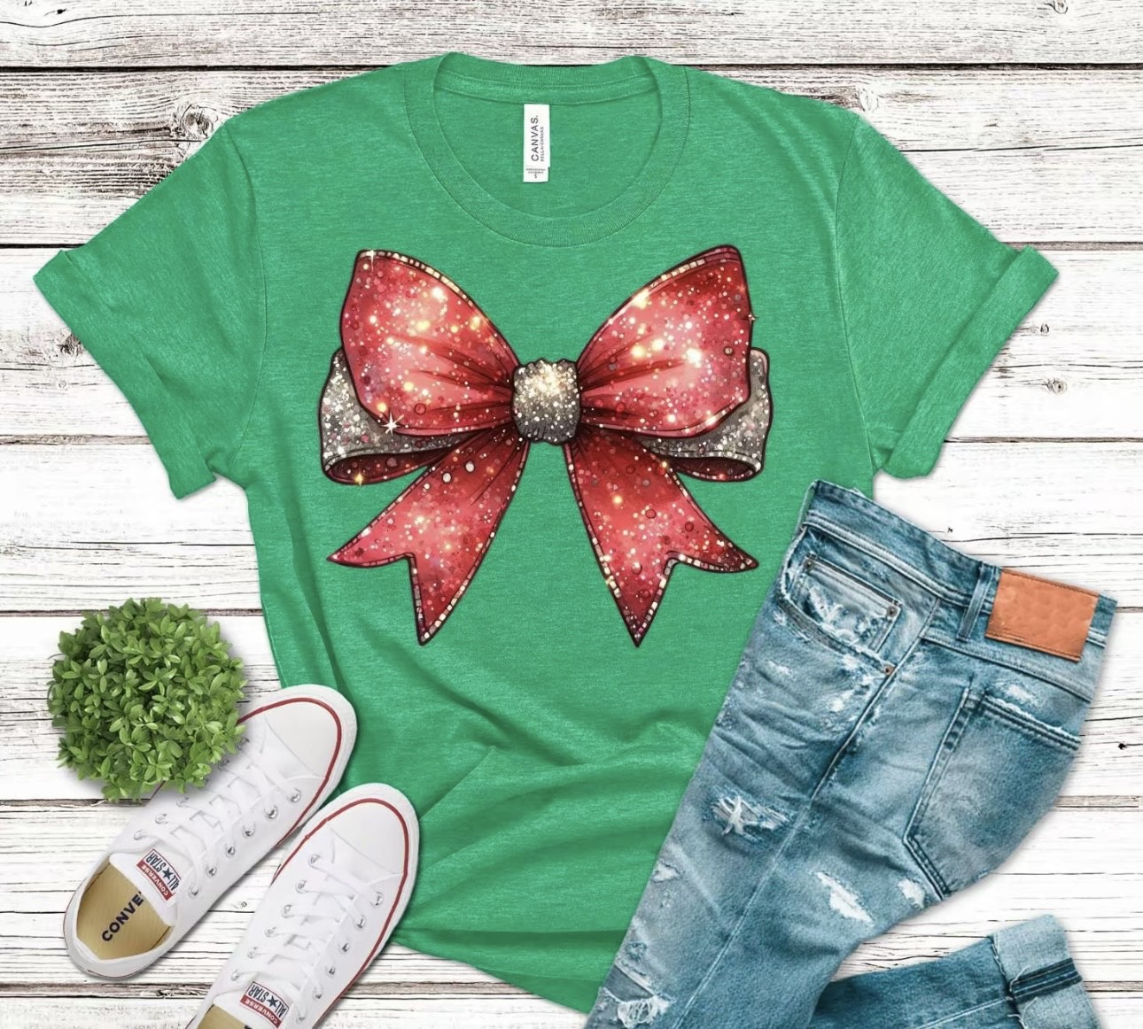 Green t-shirt with a large red bow graphic, paired with jeans and sneakers on a wooden surface.